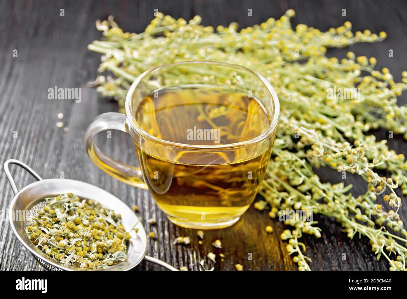 Gray wormwood herbal tea in a glass cup, fresh flowers and metal strainer with dried flowers sagebrush on wooden board background Stock Photo