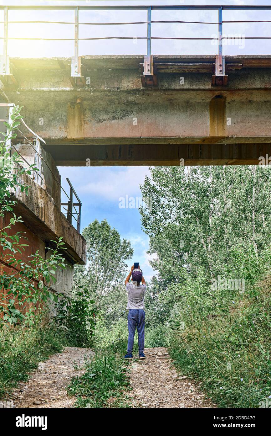 Curious boy taking pictures of the concrete support railway bridge. Iron train bridge over the river Stock Photo