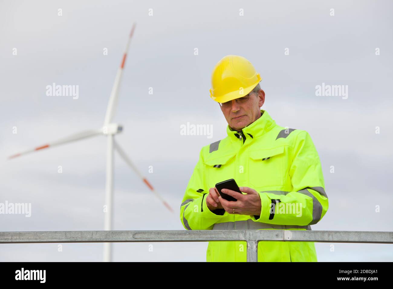 Mature engineer with protective clothing looking at a smart phone in front of a wind turbine in the blurred background Stock Photo