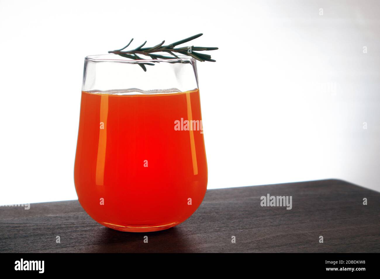 Semicircular glass of fresh orange juice with rosemary sprig on top staying on a wooden table against light background Stock Photo