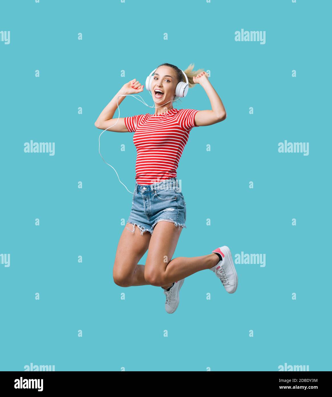 Cheerful young woman wearing headphones and listening to music, she is jumping and feeling excited Stock Photo