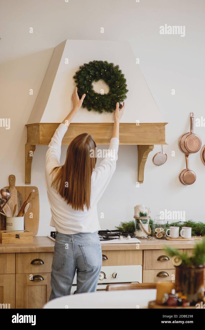 Back view of beautiful woman in white sweater hanging a Christmas wreath on her kitchen. Stock Photo