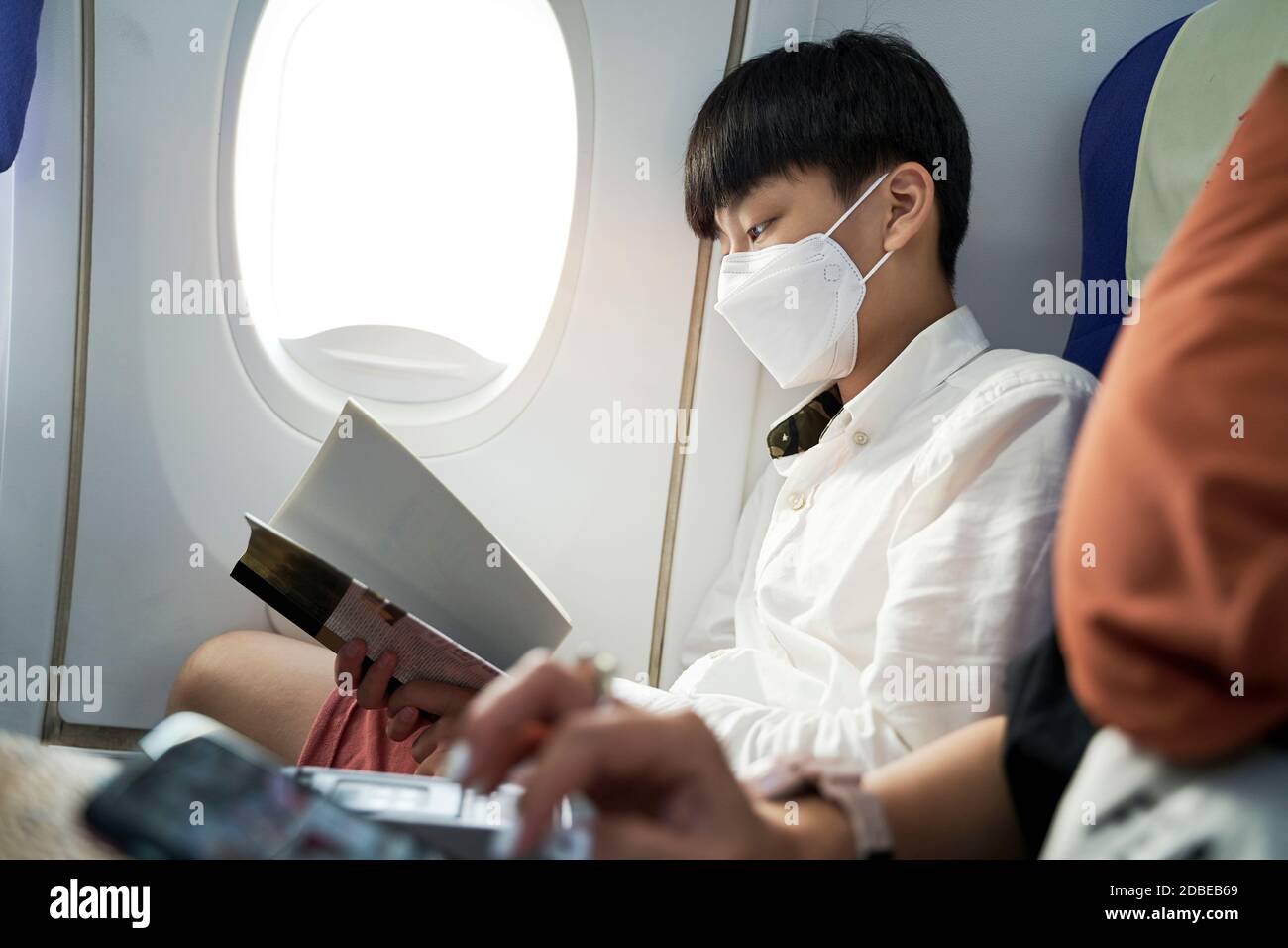 fifteen year old boy on airplane wearing face mask reading a book Stock Photo