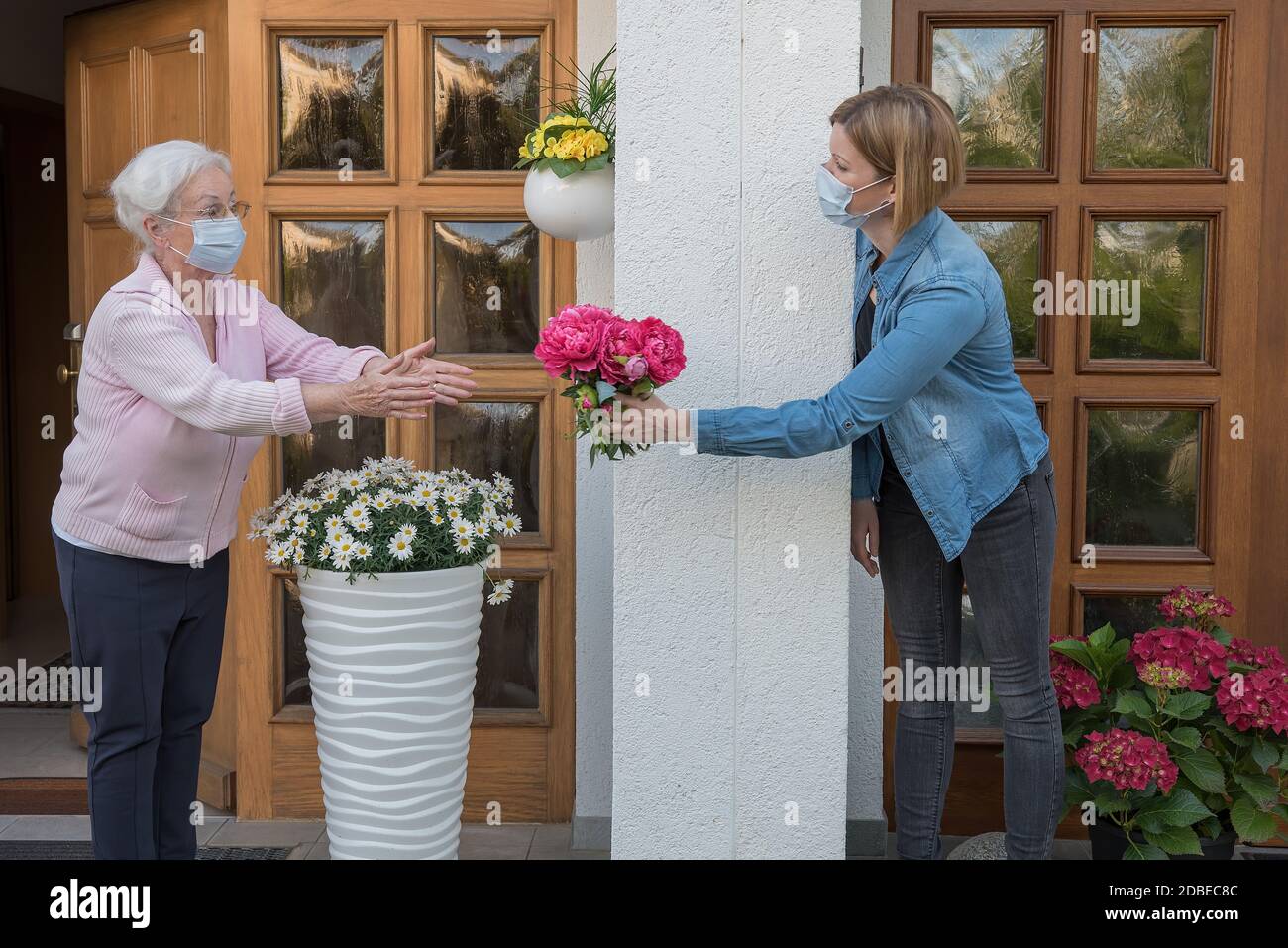 Senior woman with face mask gets flowers from young neighbor woman Stock Photo