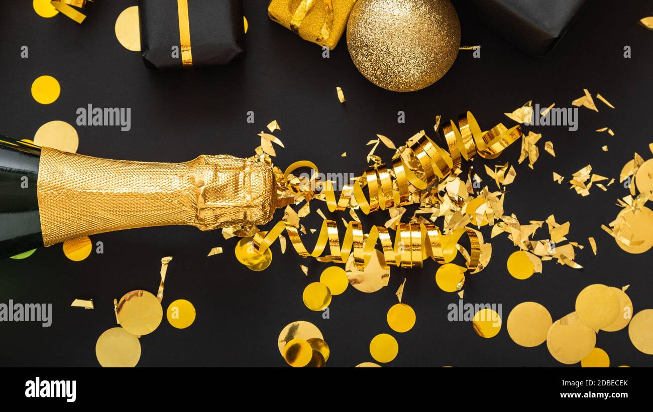 Golden bottle of champagne spills out gold sparkles in frame of gold festive Christmas decor confetti balls gifts on black background. Flat lay New Stock Photo