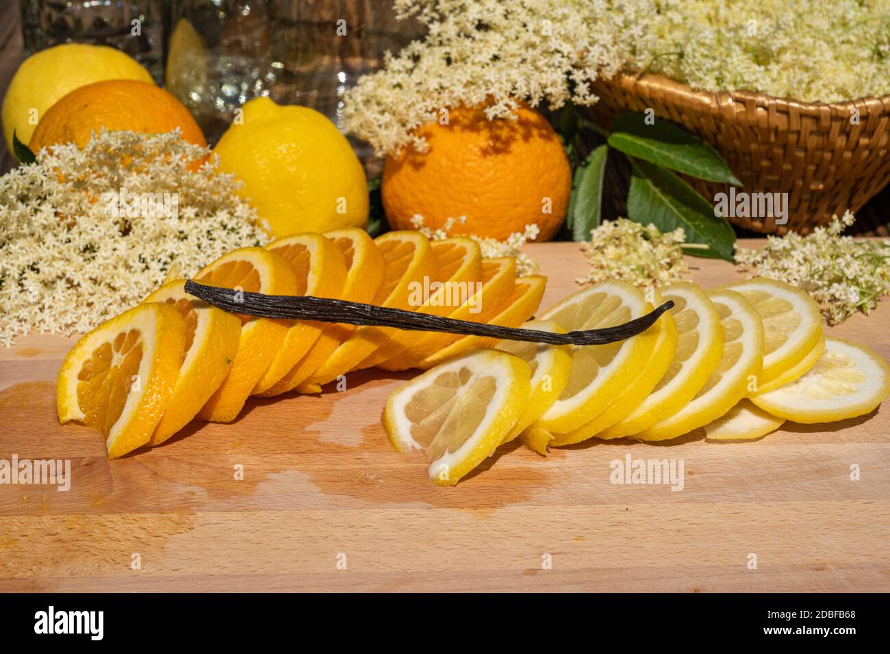 On a cutting board lie a sliced organic lemon and organic orange and a vanilla pod for the elderflower liqueur in the background are elderflowers Stock Photo