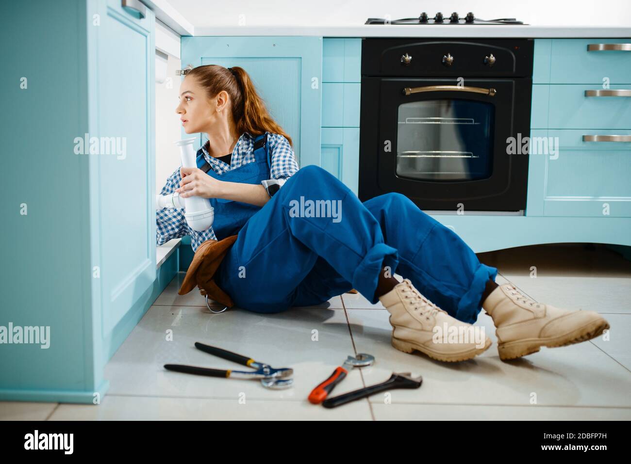 Cute female plumber in uniform holds drain pipe in the kitchen. Handywoman with toolbag repair sink, sanitary equipment service at home Stock Photo