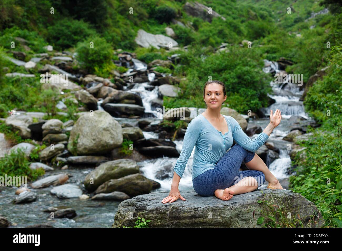 Yoga exercise outdoors - woman doing Ardha matsyendrasana asana - half spinal twist pose at tropical waterfall in Himalayas in India Stock Photo