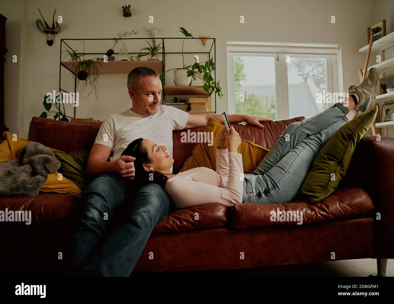 Beautiful young girlfriend using phone while relaxing on boyfriend lap on couch at home Stock Photo