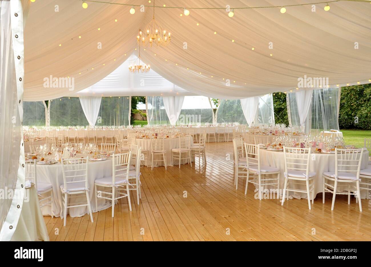 Elegant open air white marquis for a wedding venue with hardwood floor and white formal furniture decor with flowers erected in a lush green garden or Stock Photo
