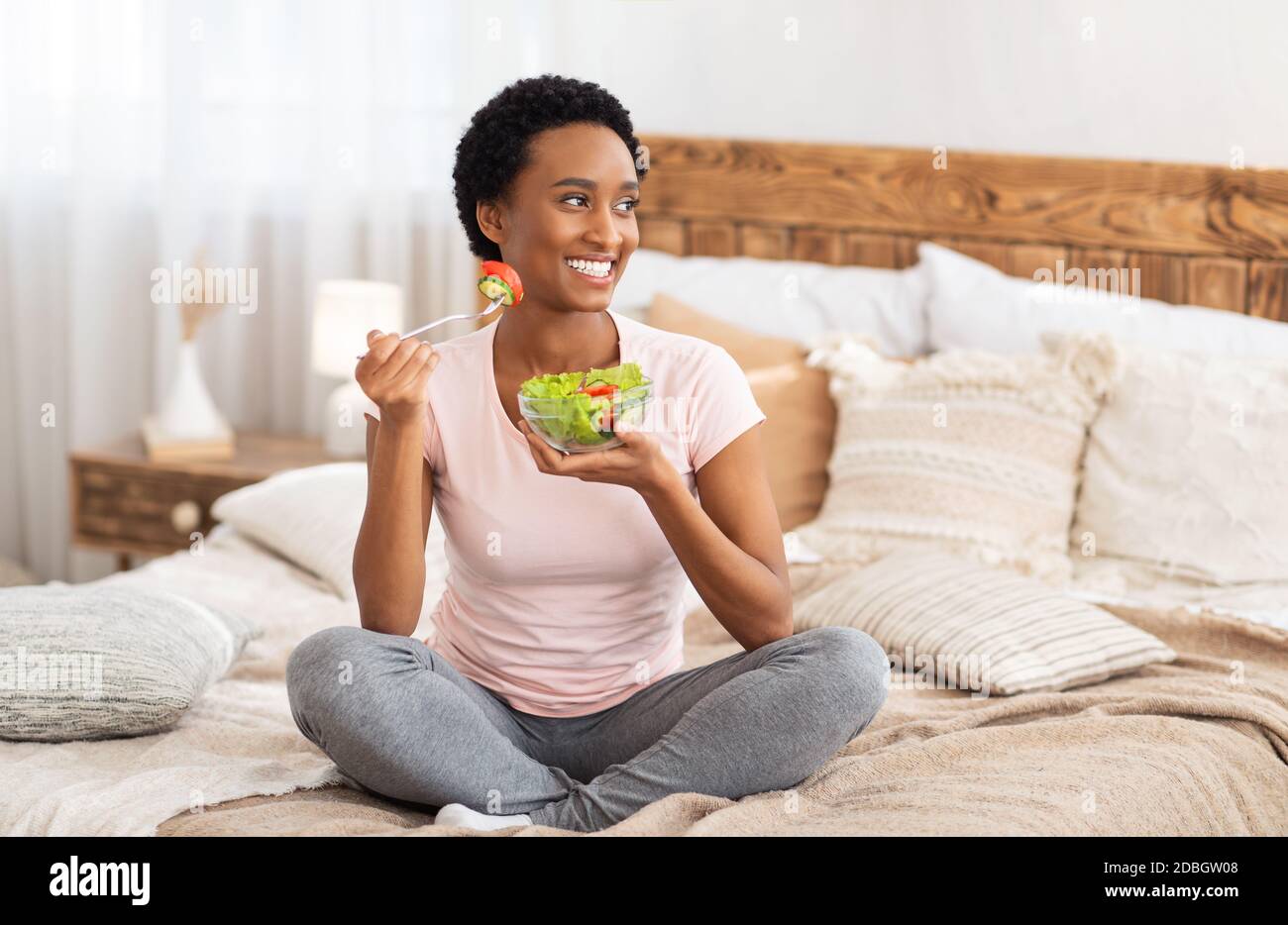 Healthy diet for weight loss concept. Happy black woman eating yummy vegetable salad on bed at home, blank space Stock Photo