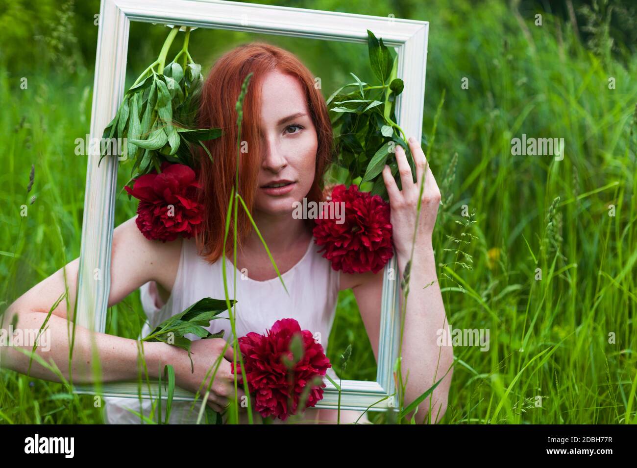 Redhead caucasian woman holding a picture frame decorated with red peony flowers. Wedding photozone concept. Stock Photo