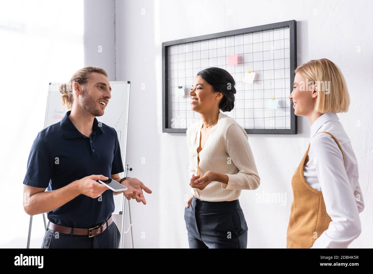 Cheerful multiethnic office workers talking while standing near mesh organizer and flipchart at workplace Stock Photo