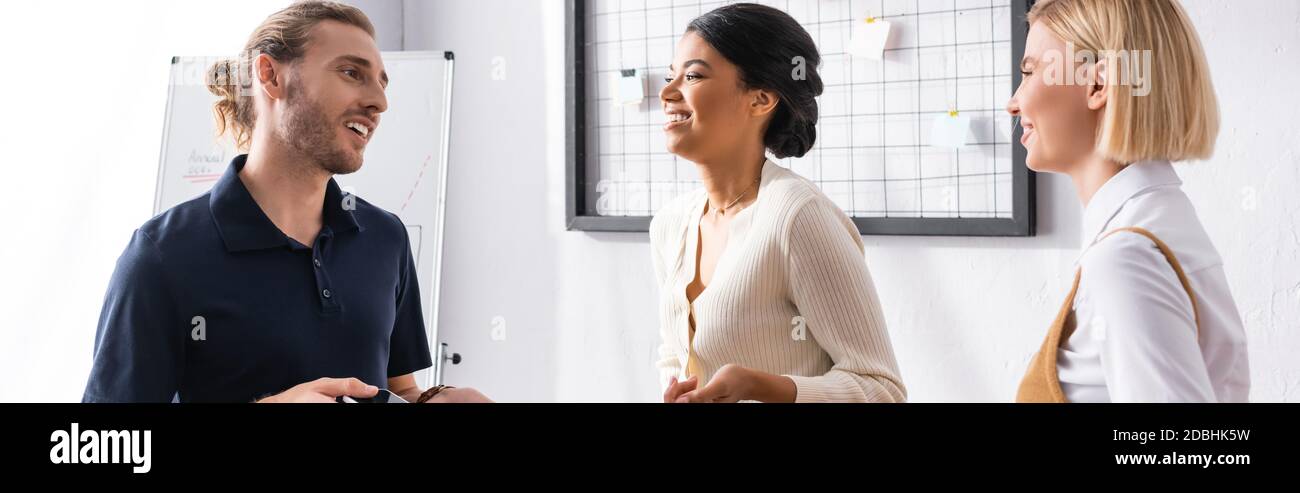 Happy multicultural office workers talking while standing near mesh organizer and flipchart at workplace, banner Stock Photo
