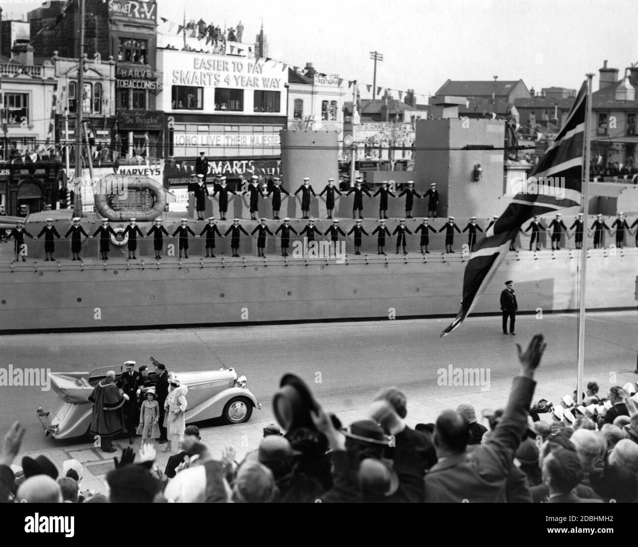 'In front of the car, from right to left: King George VI, Princess Elizabeth and Queen Elizabeth at the Royal Navy Review in Portsmouth. Young boys in sailor's uniform are standing on a decorated warship. The houses are decorated with garlands and signs saying ''Long live heir majesty''.' Stock Photo