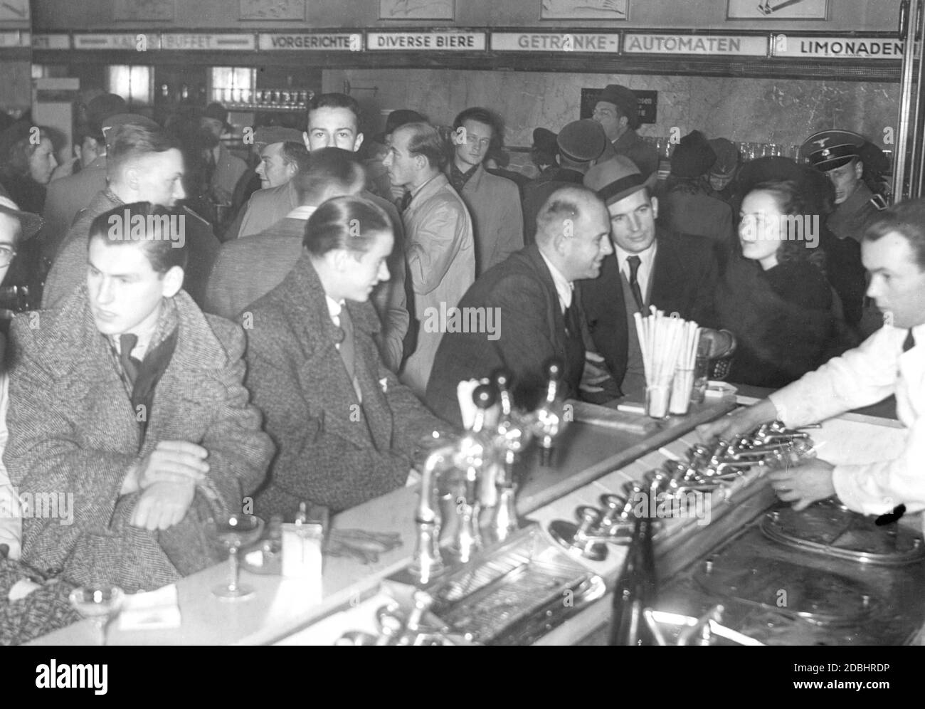 'The photo is intended to demonstrate the cheerful mood of the Berlin population in October 1939 after the outbreak of war. Many guests crowd into a bar or sit at the bar counter and chat. Among the guests are some Wehrmacht corporals, as well as a member of the SS (on the right, wearing a peaked cap, presumably an SS Scharfuehrer). The bar advertises with a large assortment: ''Cold buffet, starters, various beers, vending machines, lemonades.' Stock Photo