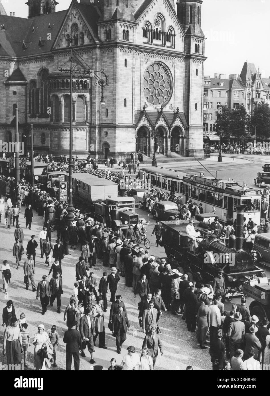 'The replica of the first German train ''Adler'' (locomotive and wagons) was pulled through Berlin to the exhibition grounds on Kaiserdamm on July 11, 1936. There it then drew visitors to the ''Deutschland'' exhibition through the open-air grounds. In this photo, the Deutsche Reichsbahn truck convoy is driving straight from Budapester Strasse to Hardenbergstrasse, passing the Kaiser Wilhelm Memorial Church, watched by spectators at the side of the road. Advertisements for ''AGB Stoffe'' and ''BMW'' hang on the lampposts.' Stock Photo