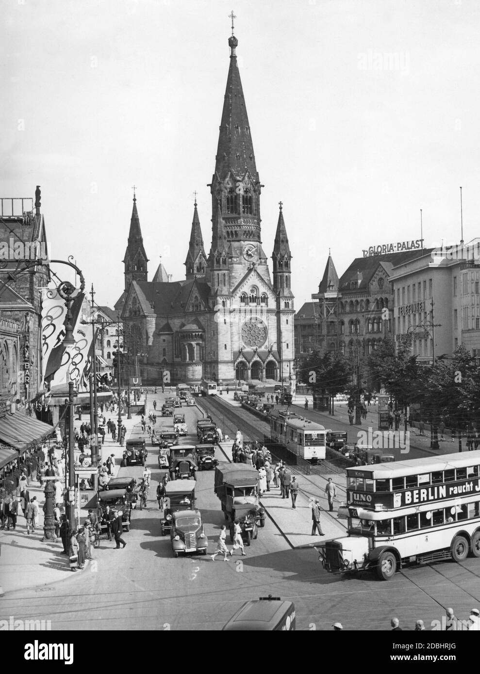 'The photograph shows the Kaiser Wilhelm Memorial Church in Berlin at the time of the Olympic Games in 1936, with the hustle and bustle of the Hardenbergstrasse, at the intersection with Joachimsthaler Strasse, in the foreground. To the right in front of the church is the Romanisches Haus with the Gloria-Palast, next to it is a slogan ''Telefunken - Die Deutsche Weltmarke'' (''Telefunken - The German World Brand''). On the left is the Haus Germania and the Ufa-Palast am Zoo. A double-decker bus of line 20 to ''Knie'' with the advertisement ''Berlin raucht JUNO'' (''Berlin smokes JUNO'') Stock Photo