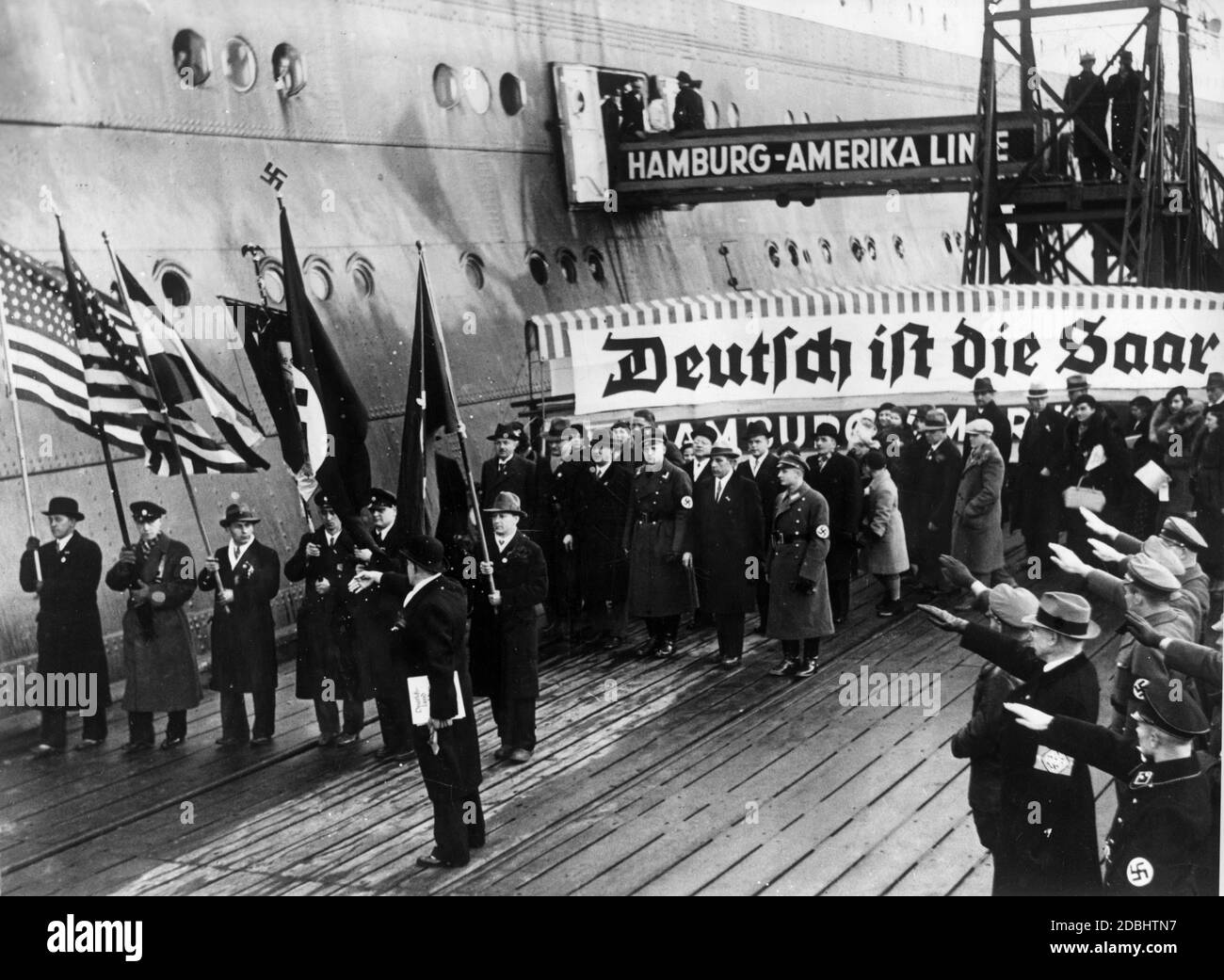 'The passenger steamer ''Deutschland'' of the Hamburg - Amerika Line arrived in Cuxhaven with 358 Saar Germans from America who came to participate in the referendum on the annexation of the Saar region to the German Reich. At the landing pier they are received by National Socialist organizations. The picture shows the people of Saarland with their association banner (''The Saar is German!''), American national flags and swastika flags.' Stock Photo