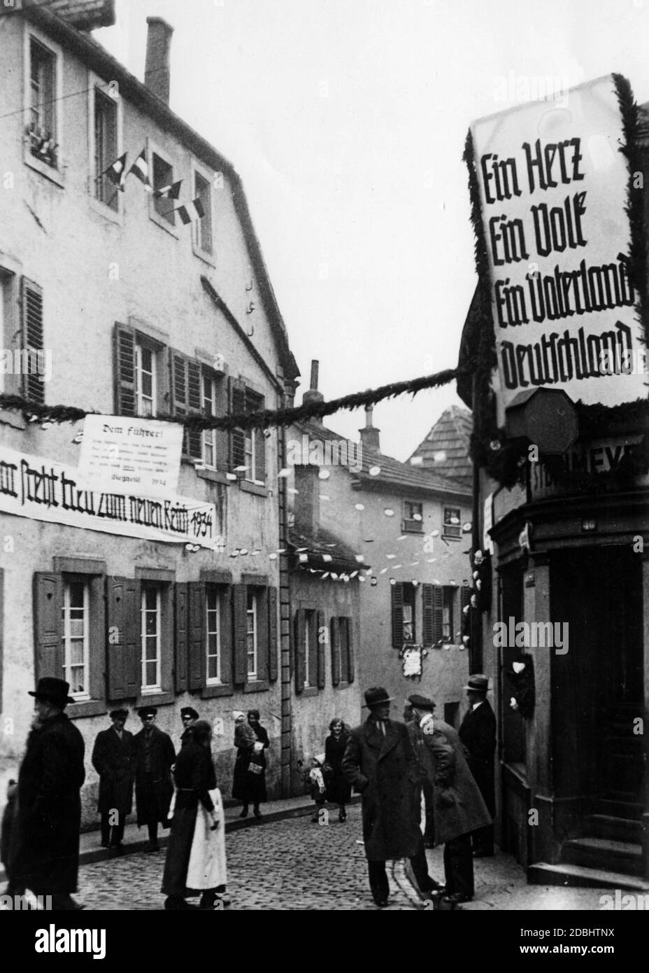 'The Altneugasse of Saarbruecken is festively decorated with swastika flags, imperial flags and banners to celebrate the first anniversary of the ''seizure of power''. Left: ''Saar strives faithfully to the new Reich 1934''. In the middle a text '' To the Fuehrer!''. On the right the slogan: ''One Heart. One Nation. One Fatherland. Germany''.' Stock Photo