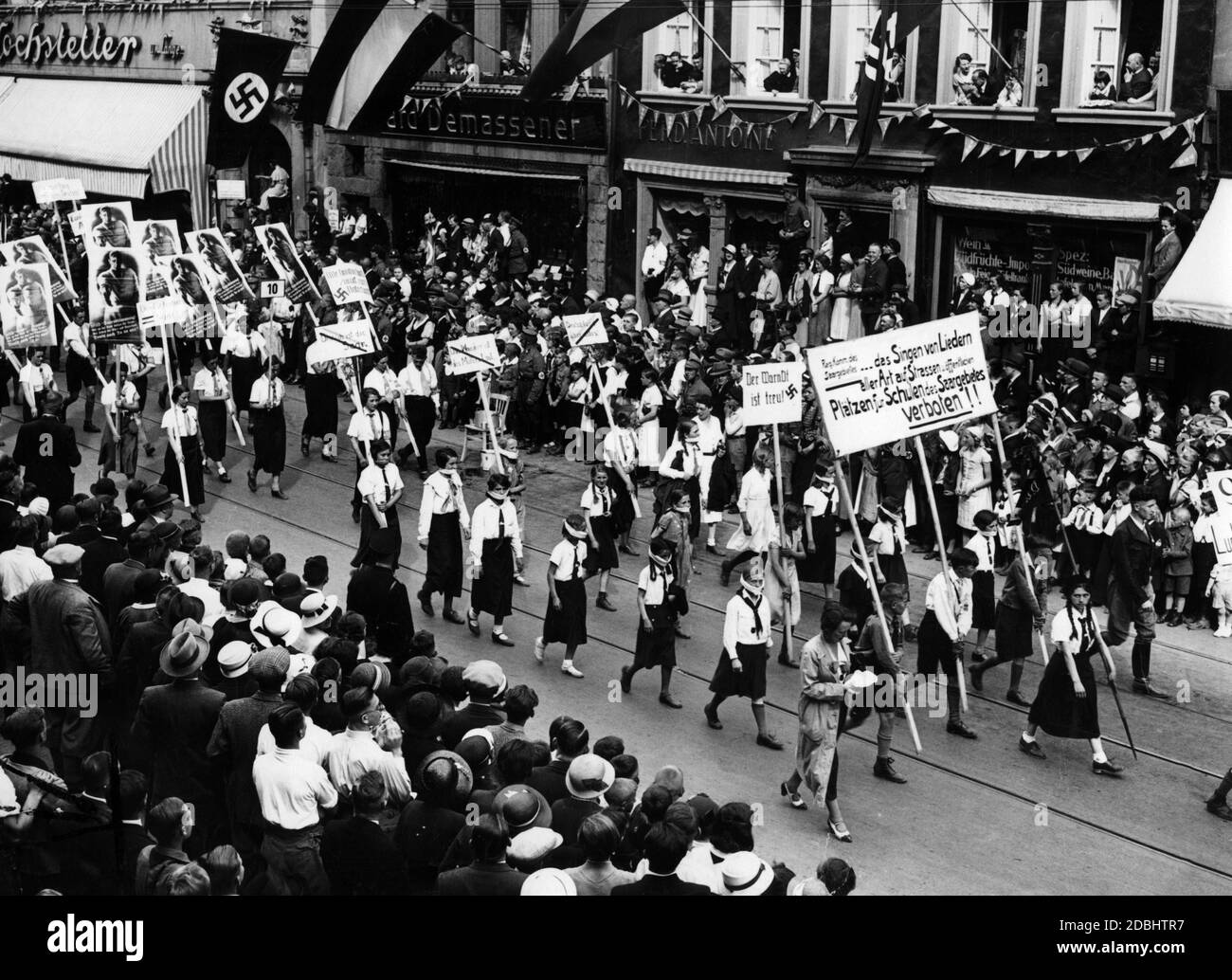 'During the VDA conference at the Rhine and Moselle, a parade is taking place in the streets of Trier. Children from the Saarland illustrate the Saar government's ban on singing songs in public streets and squares with banners and posters and a gag around the mouth. The sign in front says: Government Commission of the Saar region... the singing of songs of all kinds is forbidden for schools of the Saar region in streets and public places!'' On the sign behind: ''The Warndt is loyal!''' Stock Photo