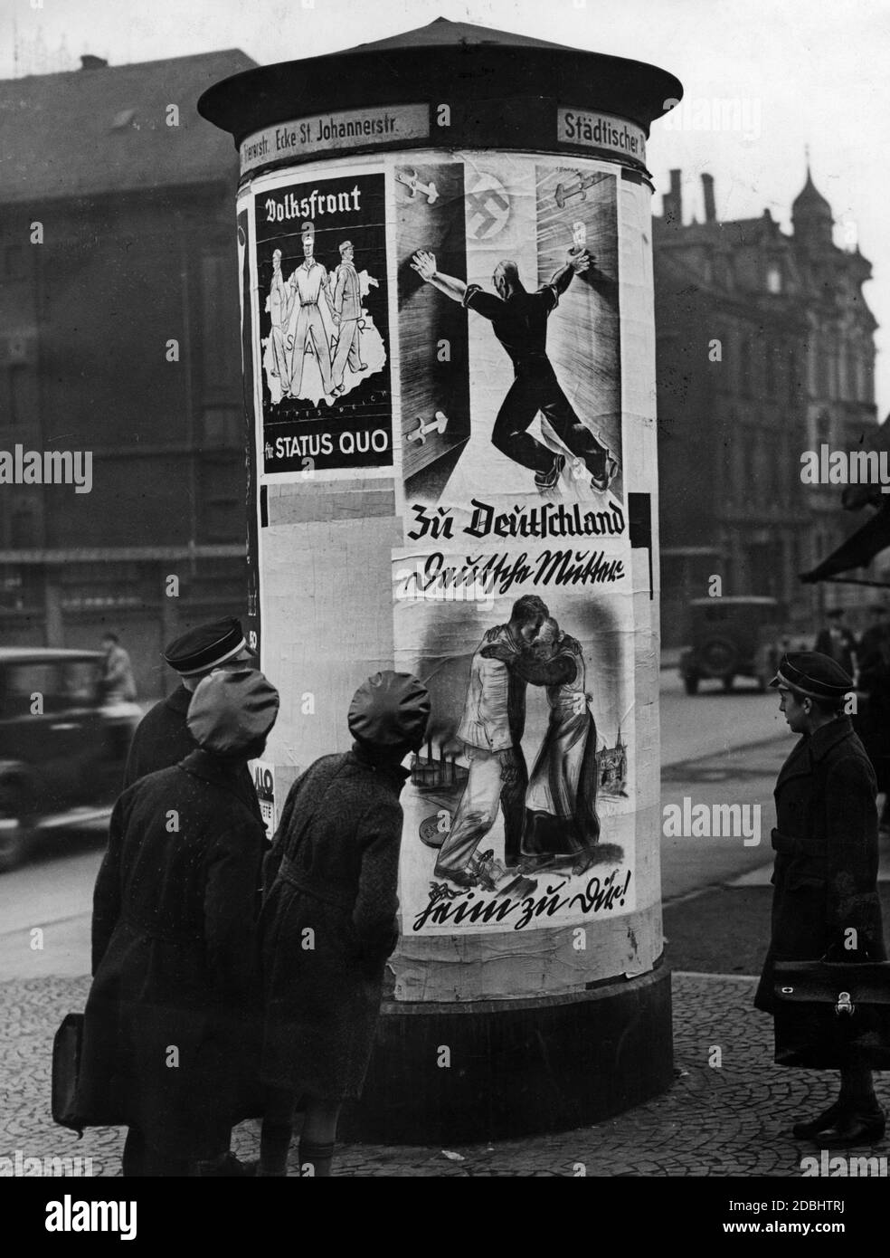 'At one of the advertising pillars on the corner of St. Johannerstrasse in Saarbruecken, passers-by look at election posters for the referendum on the annexation of the Saar region to the German Reich on 13.01.1935. On the left, the election poster of the separatist ''Volksfront''. Three men who are ready to fight back stand arm in arm on a map of the Saar area against the Third Reich. Caption: ''For Status Quo''. On the right the posters of the Deutsche Front. Above: ''To Germany''. A man opens the gate to the Third Reich, symbolised by the swastika. Below: ''German Mother. Home to you!''' Stock Photo