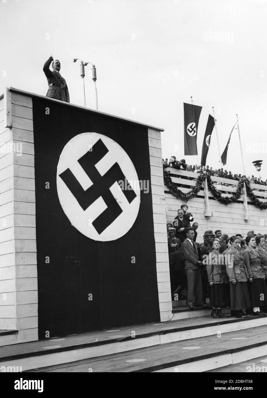 Adolf HItler holds a speech during the parade of the Nazi Labor Service from the swastika-decorated speaker's stand on Zeppelin Field on the Nuremberg Reich Party Rally Grounds. To the right of the swastika flag hangs the flag of the RAD. Below the tribune are several women of the Labour Service. Stock Photo