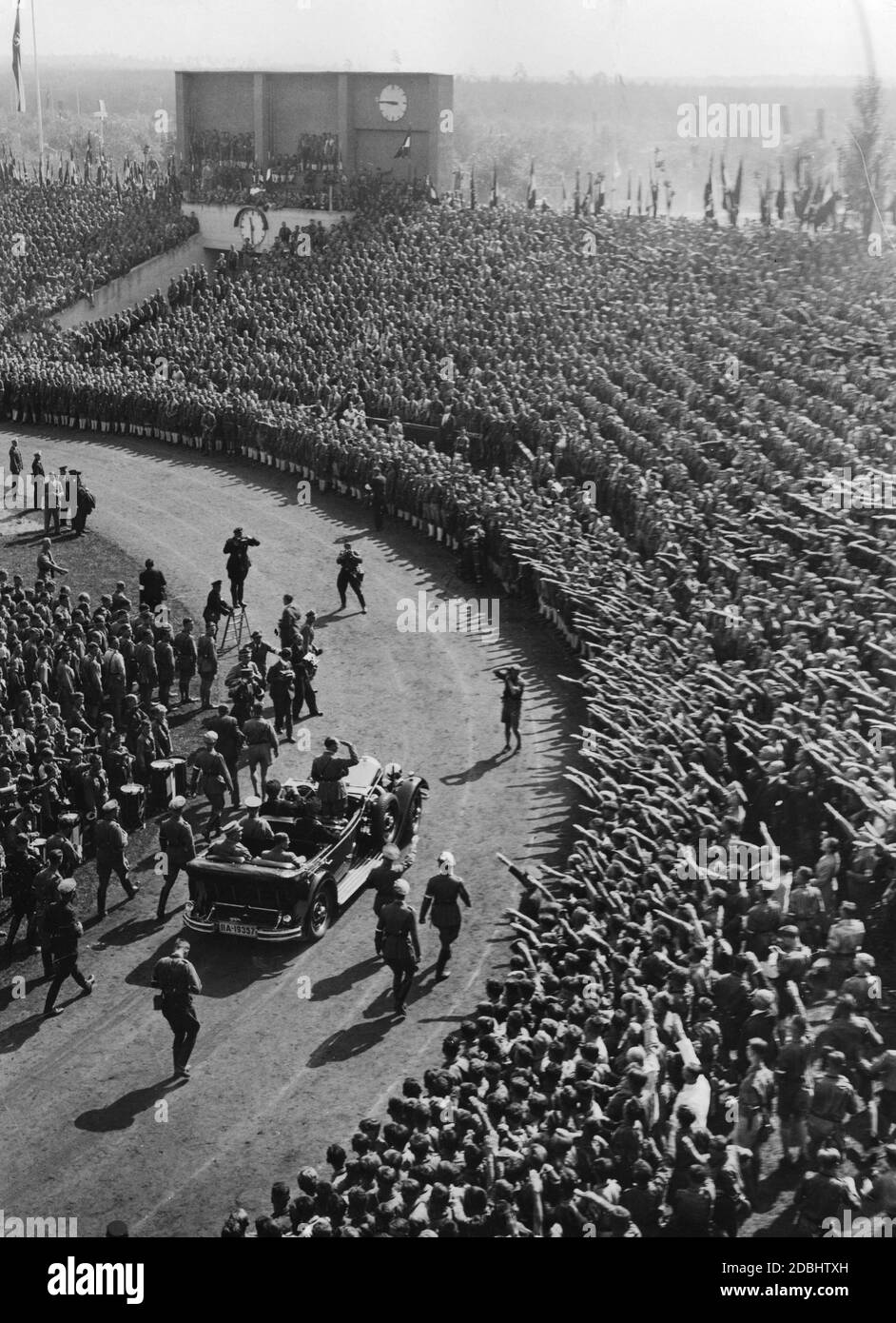 Adolf Hitler, standing in a Mercedes, drives around in the Municipal Stadium and greets the participants of the Hitler Youth at their rally during the Nazi Party Congress in Nuremberg. On the back seat on the left, Adjutant Wilhelm Brueckner, on the right, Baldur von Schirach. On the left is a drum corps of the Hitler Youth. Stock Photo