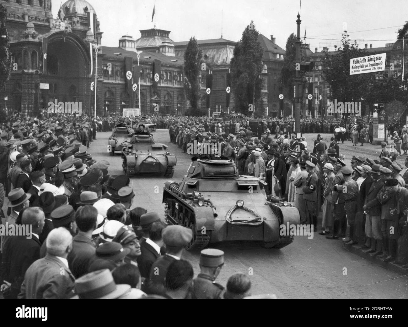 'Before the ''Day of the Wehrmacht'' at the Nazi Party Congress in Nuremberg a column of new tanks (Panzerkampfwagen I) rolls through the city centre, here at the Bahnhofsplatz (in the background is the station building decorated with swastika flags). A sign on the right points to the ''Koenigstor stop, boarding point to Zeppelinwiese at Marientor'' in a two minutes walk. The tanks still bear the ''colourful paint'' as camouflage, which was common until the mid-30s, recognizable by the fleck patterns.' Stock Photo