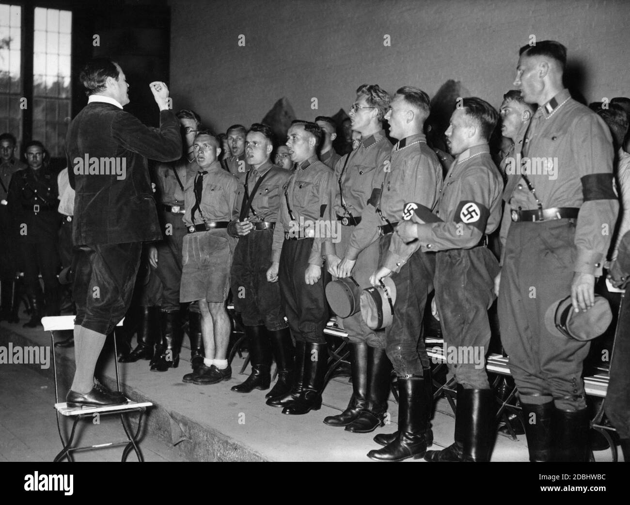 'Young SA men singing together on a small stage. The choir is conducted by Dr. Hensel Janiczek. They sing at the first Reichssingwoche (''Reich Singing Week'') in the festival hall of the airship airport Potsdam. The event is organized by the National Socialist Community ''Kraft durch Freude'' (Strength through Joy) and the German Student Union.' Stock Photo