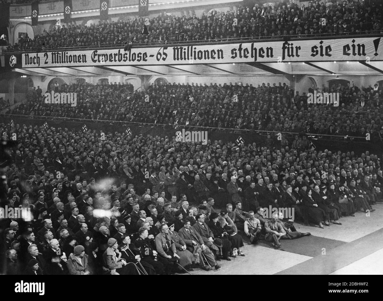 'View of the audience during a speech by Joseph Goebbels at the Berlin Sportpalast. Some SA members are sitting on the floor. A banner reads ''2 million unemployed - 65 million still stand up for them!''' Stock Photo