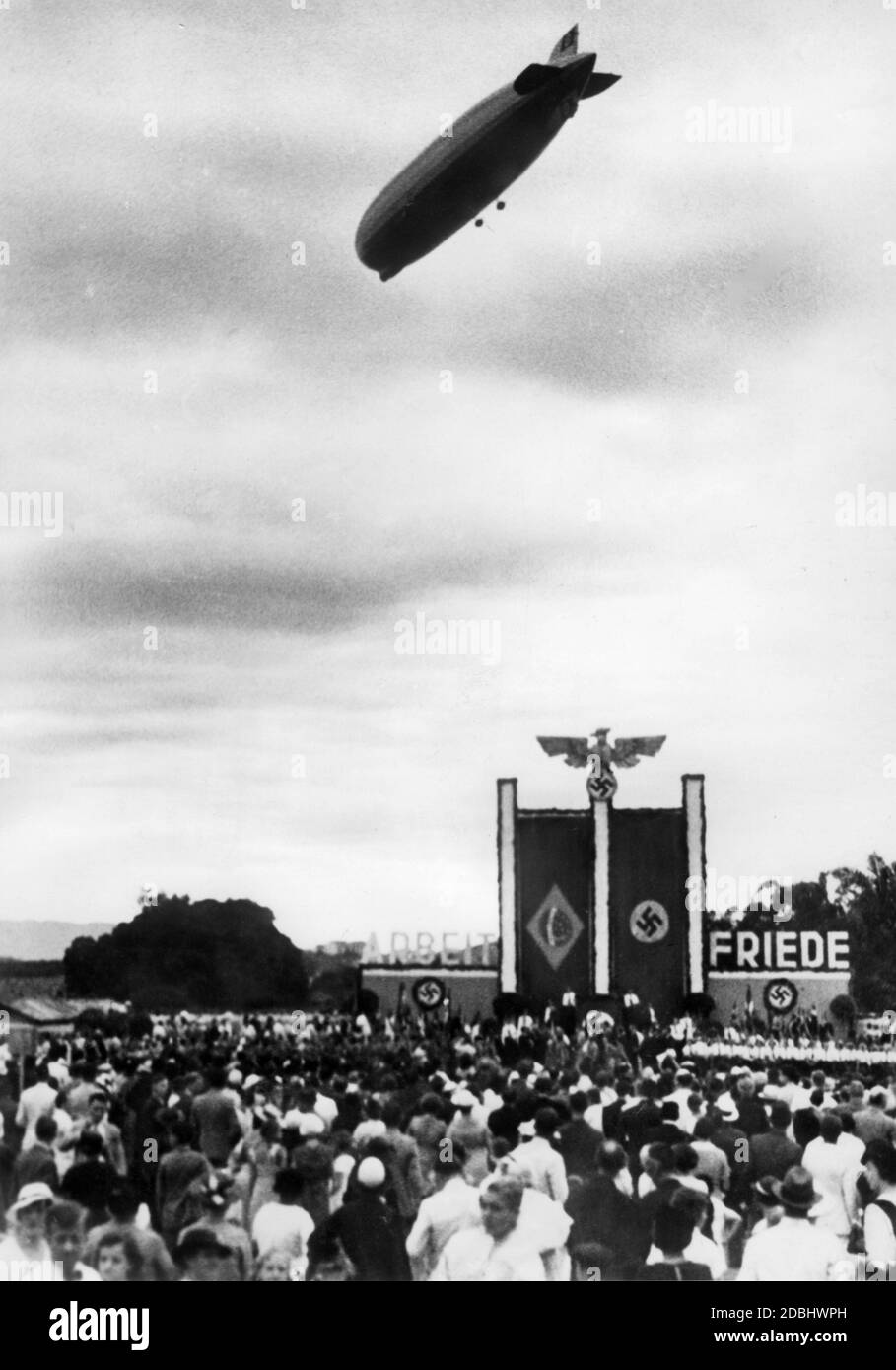 'The LZ 127 ''Graf Zeppelin'' is welcomed by the German colony in Rio de Janeiro, who celebrate the ''National Day of the German People'' for the third time on the square of the Deutscher Turn und Sportverein. On the grandstand, which is decorated with the national flag of Brazil as well as the swastika flag and the symbols of sovereignty of the German Empire, the words ''Work'' and ''Peace'' are written with oversized letters.' Stock Photo