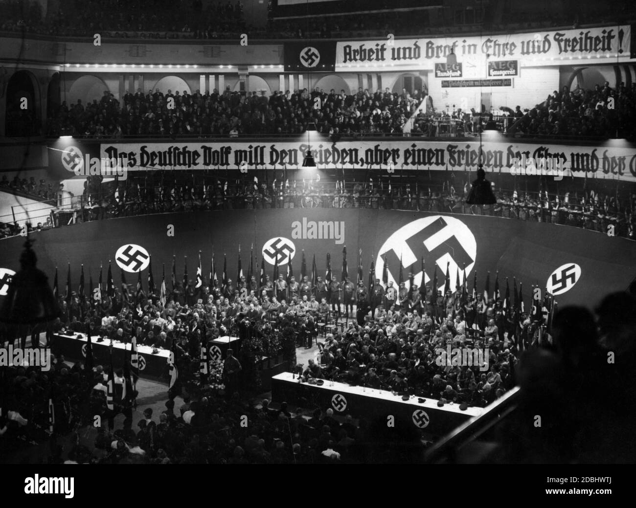 'Propaganda banners during an event of the NSDAP. ''Arbeit und Brot in Ehre und Freiheit'' (Work and bread in honor and freedom). Under the banner hangs an advertisement of the company AEG Haushaltgeraet with the slogan ''Im Gleichschritt mit unserer Zeit'' (Marching in step with our time). Underneath it is a banner with the slogan ''Das Deutsche Volk will den Frieden, aber ein Frieden der Ehre und der Gleichberechtigung'' (The German people want peace, but a peace of honour and equality).' Stock Photo