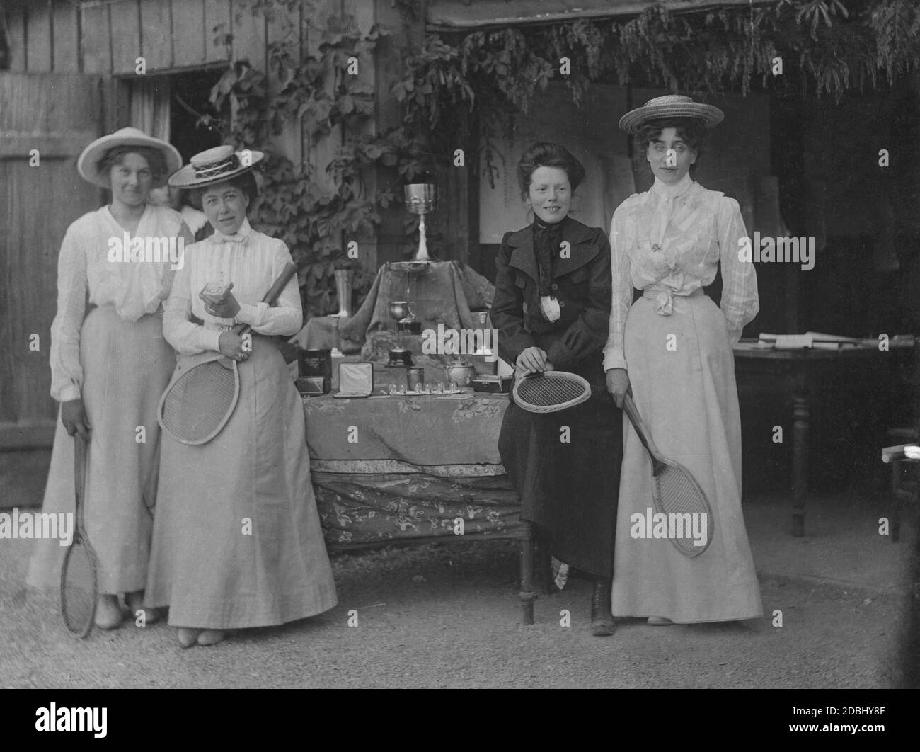 The four winners of the international Lawn Tennis Tournament stand with their rackets in front of the table with the winnings. They are wearing fancy clothes and hats. There are cups to win. From left: Mrs. Kaufmann, Mrs. Mellinger, Baroness Ungelter and Mrs. Sinner. Stock Photo