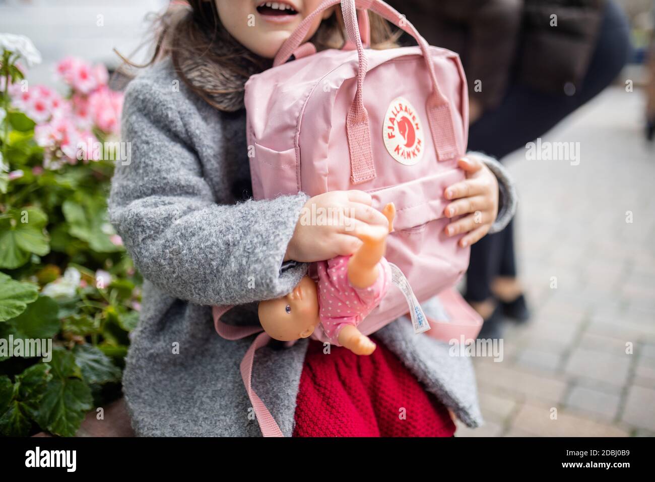 Little girl with flowers behind her hugging a pink backpack and small doll Stock Photo