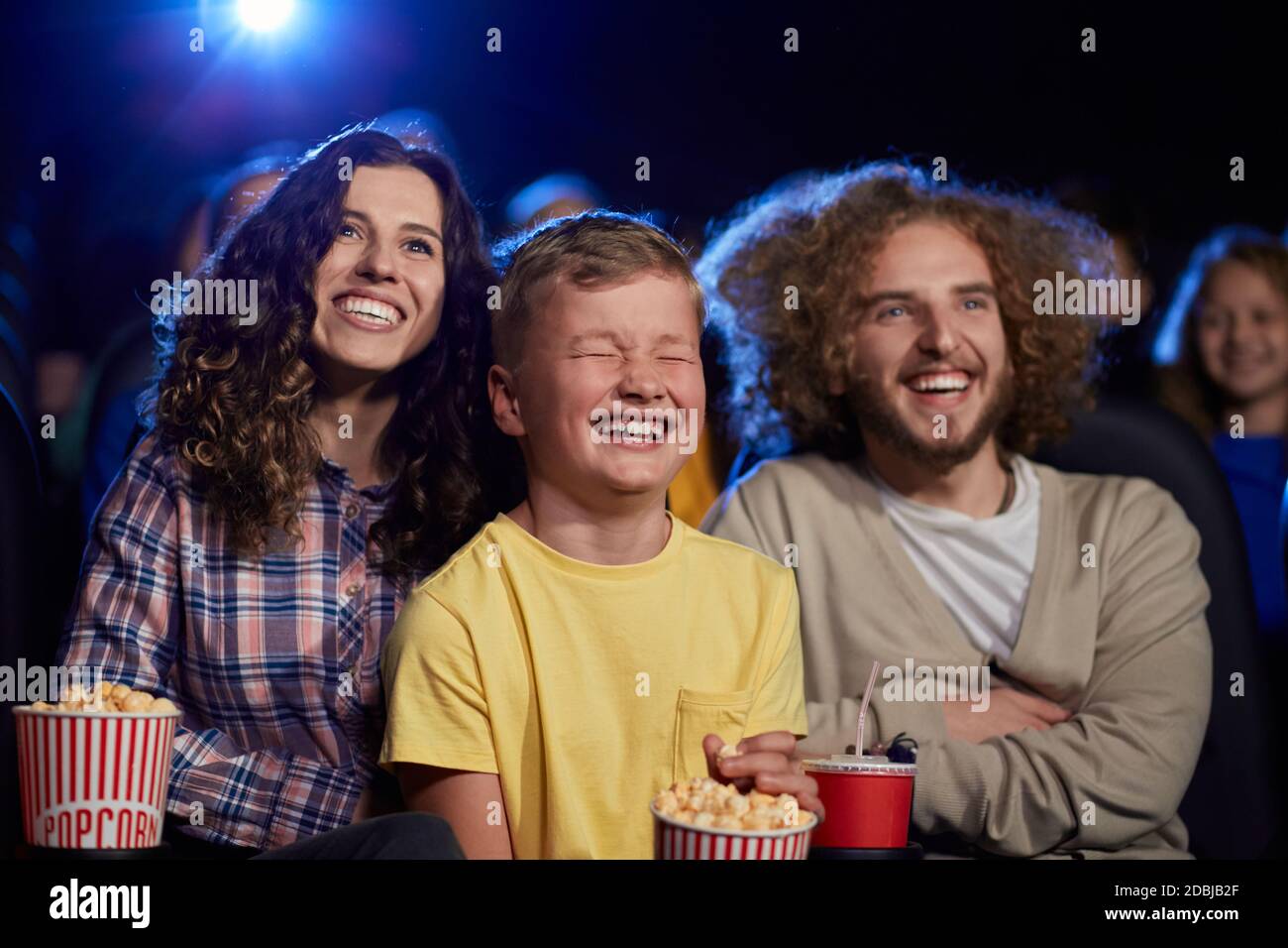 Selective focus of parents with curly hair enjoying time with son holding sparkling drink in cinema. Happy young family sitting in cinema, kid laughing with closed eyes, watching funny cartoon. Stock Photo