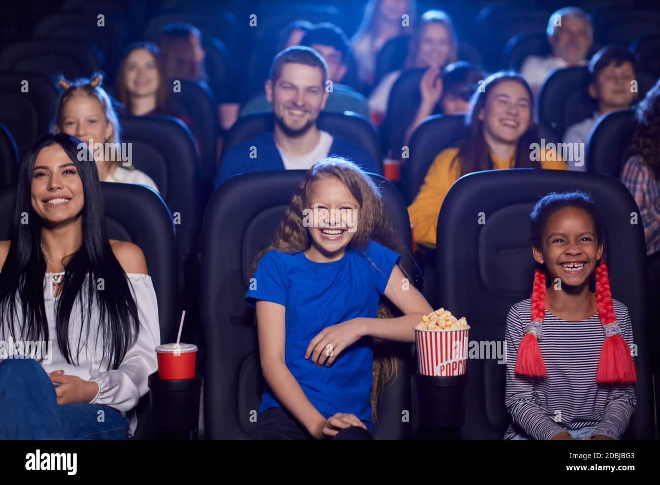 Cheerful kids enjoying time together holding sparkling drink, young multiracial audience on background. Happy little friends sitting in cinema, watching funny cartoon. Entertainment concept. Stock Photo