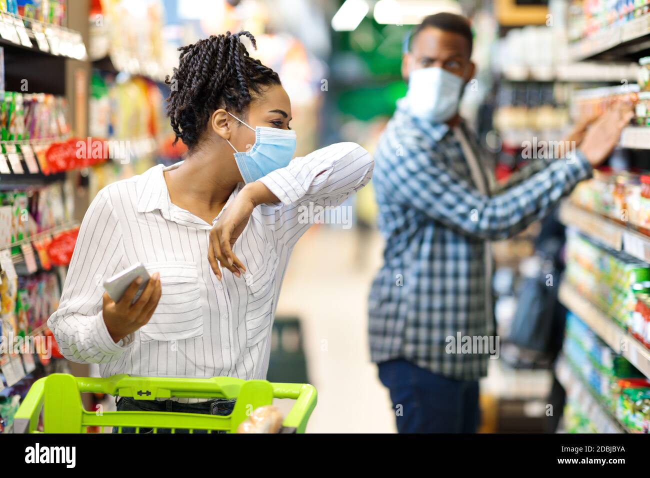 Sick African Woman Coughing In Elbow Wearing Mask In Supermarket Stock Photo