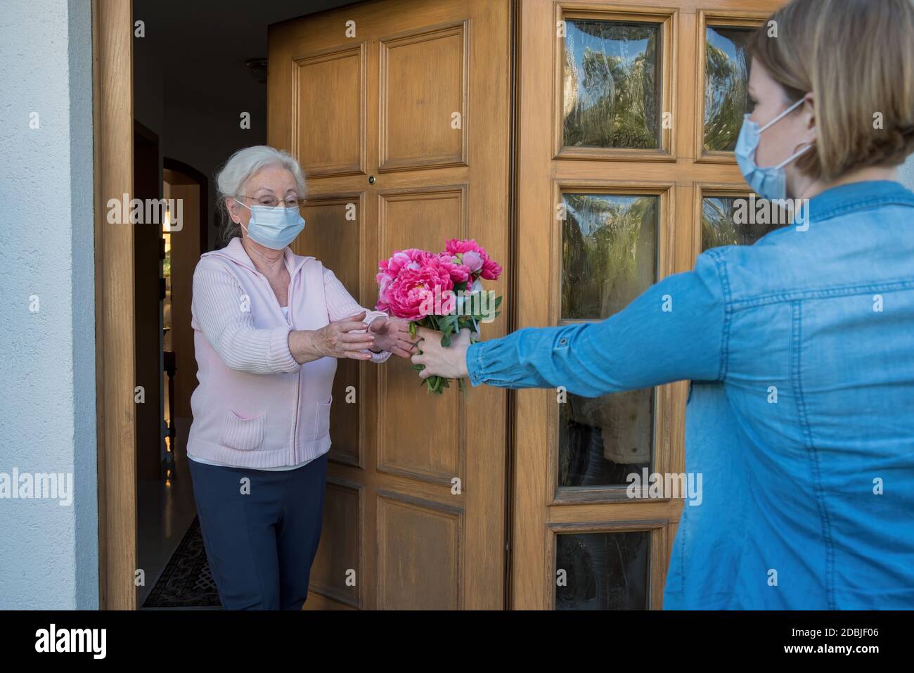 Senior woman with face mask gets flowers at the house door from neighbor woman Stock Photo