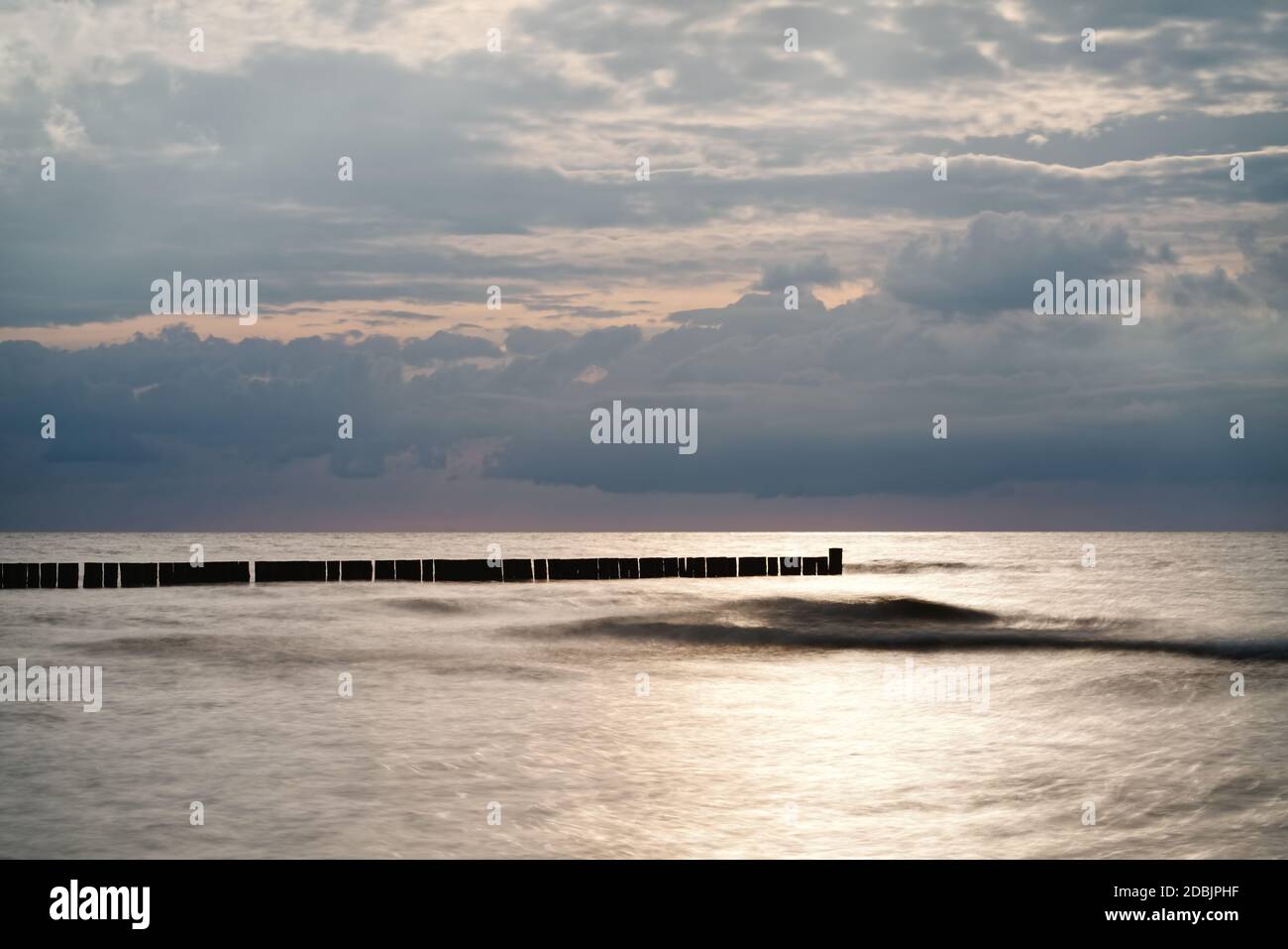 Quiet beach shot of the Baltic Sea coast with a row of groynes parallel to the horizon, in front of it a prominent wave, the water surface is smoothed Stock Photo