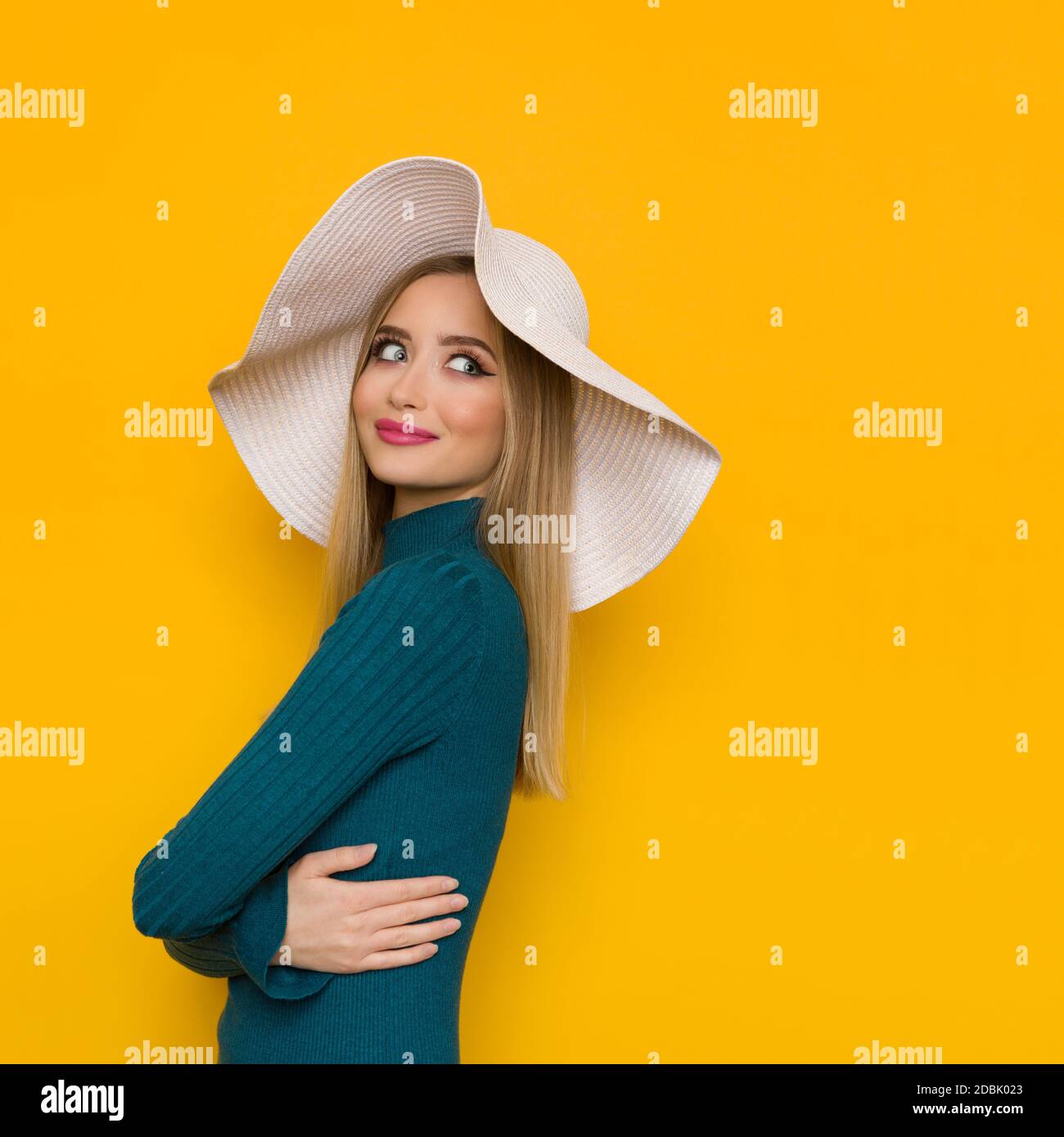 Beautiful young woman is posing in white sun hat and teal sweater, holding arms crossed and looking away over the shoulder. Side view. Waist up studio Stock Photo