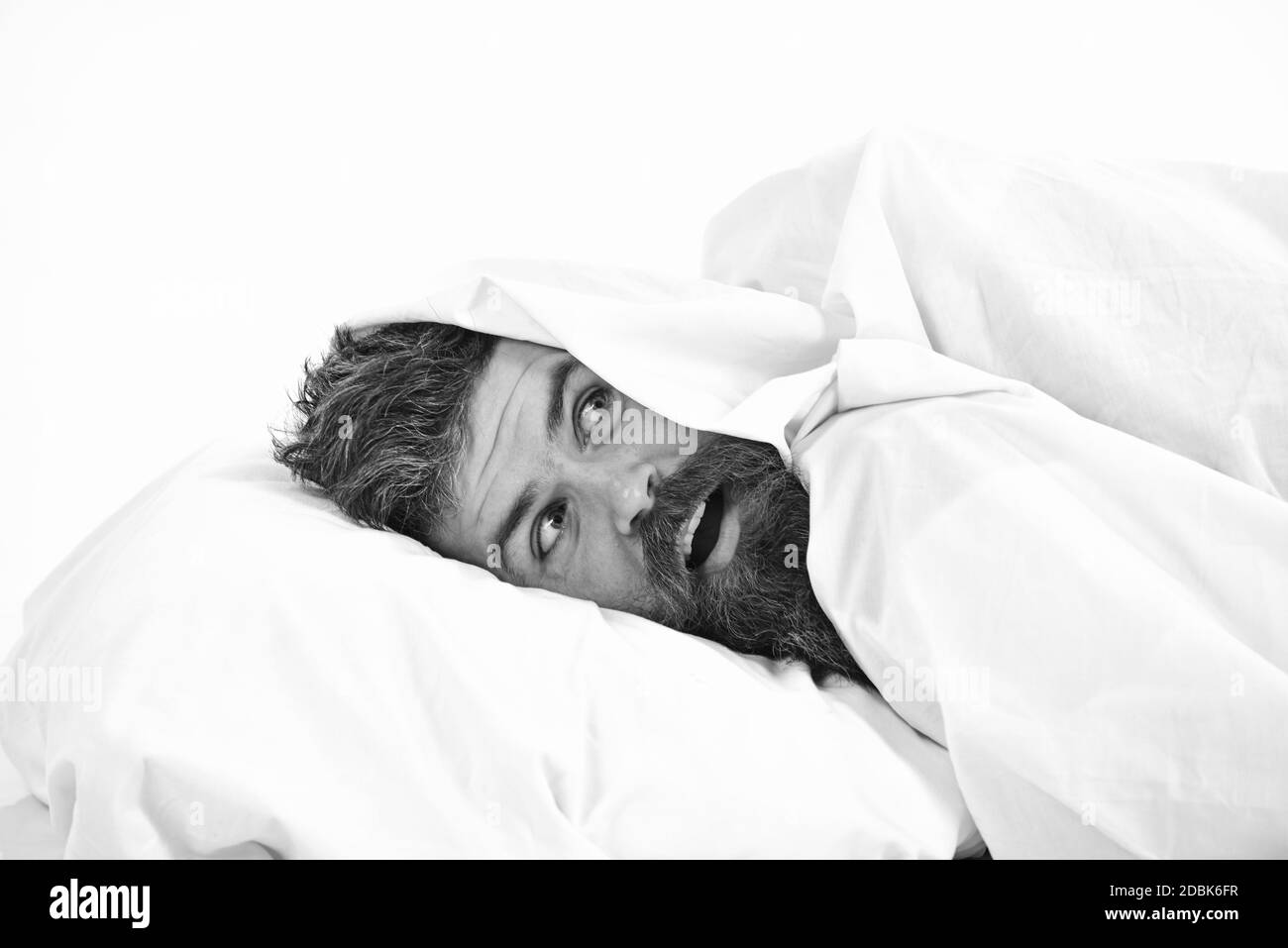Man with beard and mustache hiding in bed under blanket at home. Guy with surprised face lay under white blanket. Stock Photo