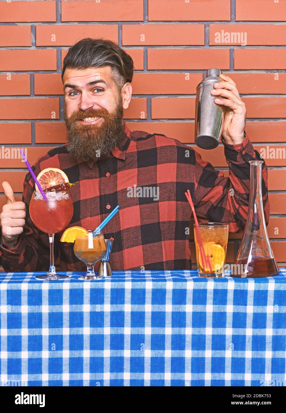 Alcoholic drinks concept. Man in checkered shirt on brick wall background prepares drinks. Barman with long beard and mustache and stylish hair on happy face holding shaker, makes alcoholic cocktail. Stock Photo