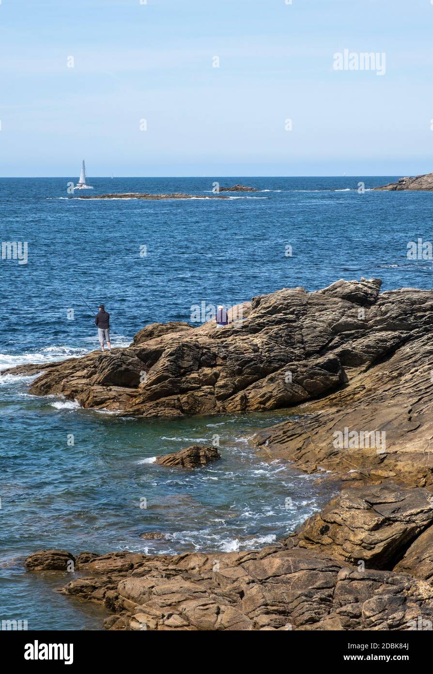 Man fishing on the rocks with a female companion and a yacht sailing past, at The Cote Sauvage on The Quiberon Peninsula, Brittany, France. Stock Photo