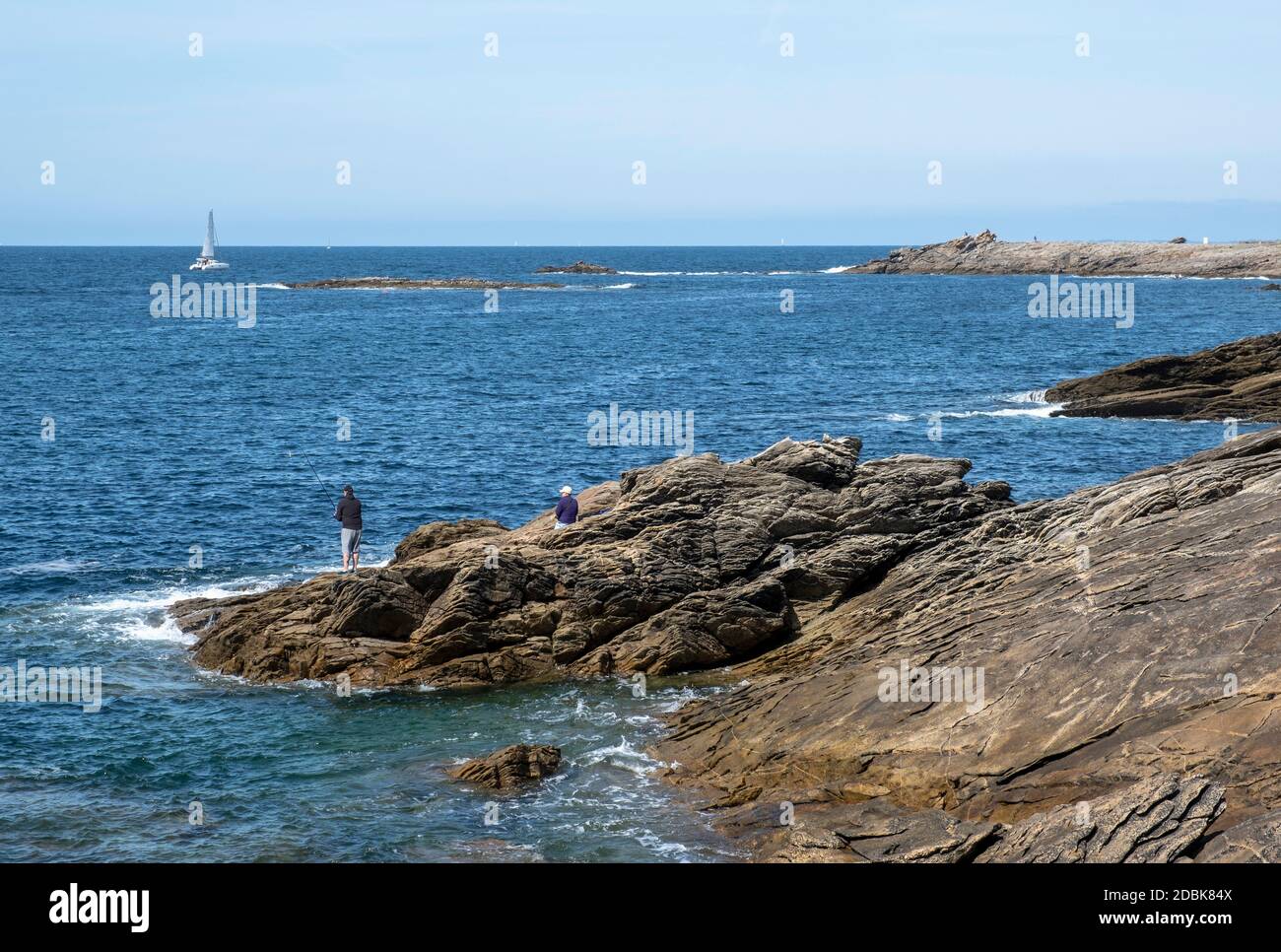 Man fishing on the rocks with a female companion and a yacht sailing past, at The Cote Sauvage on The Quiberon Peninsula, Brittany, France. Stock Photo
