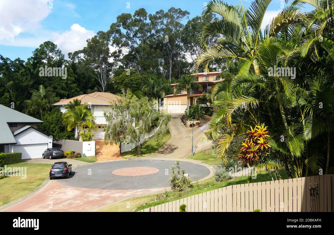 Cul-de-sac in Queensland Australia suburb with houses up on hills with steep driveways and tall gum trees behind and tropical landscaping Stock Photo