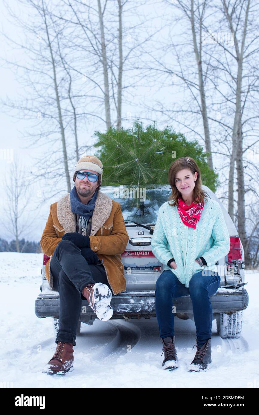 Portrait during winter of a young couple sitting on the tailgate of their small car with a christmas tree on top. Sandpoint, IDaho. Stock Photo