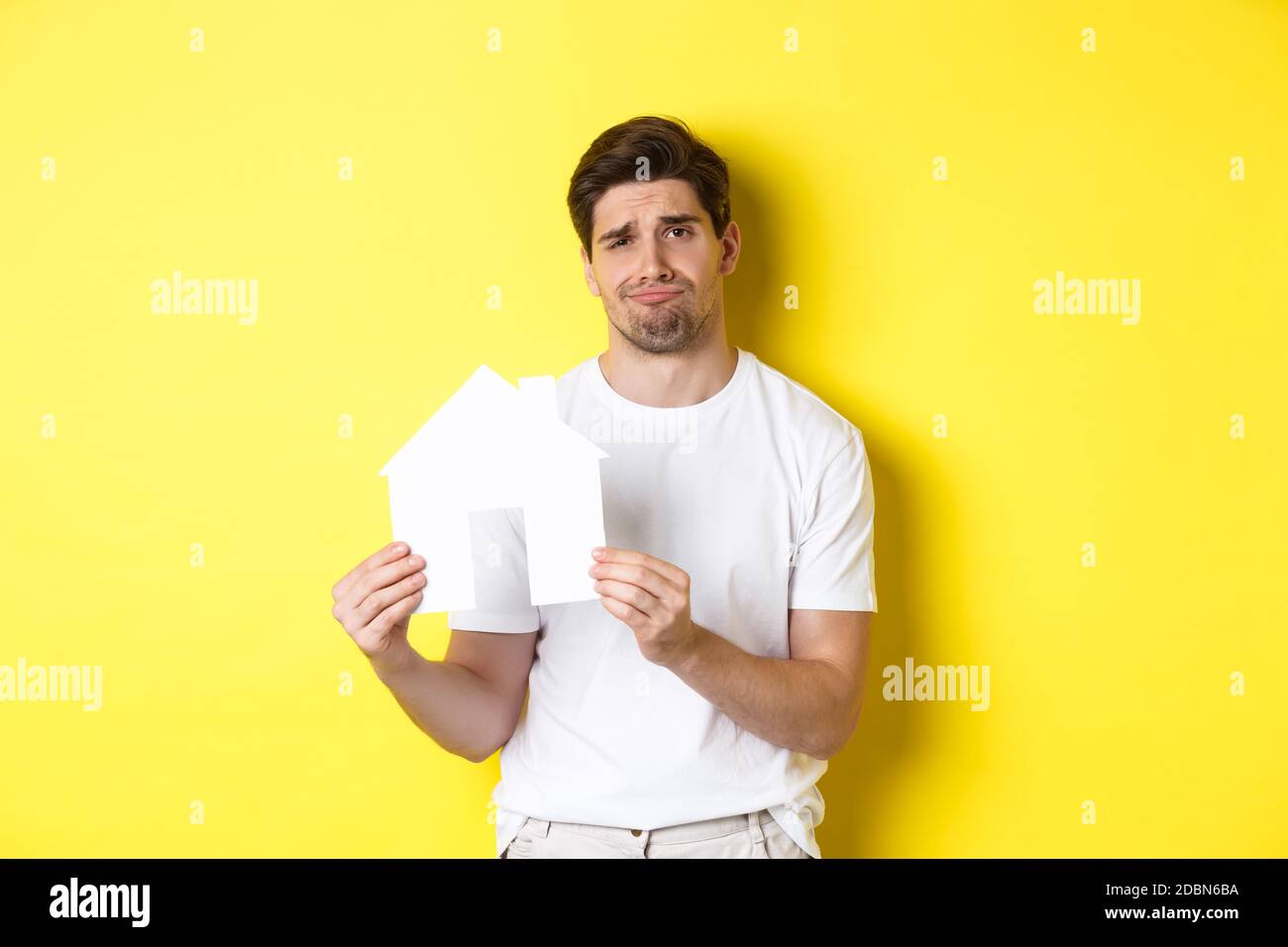 Real estate concept. Displeased young man showing paper house model and grimacing upset, standing over yellow background Stock Photo