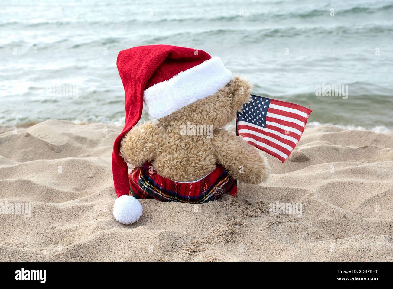 back view of brown teddy bear with Santa Claus hat and American flag sitting on beach sand looking at the ocean Stock Photo