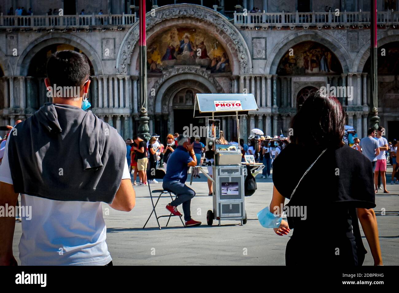 Venice tourists with surgical masks walk towards St Mark's Basilica and a man selling professional photographs during coronavirus crisis in Italy. Stock Photo